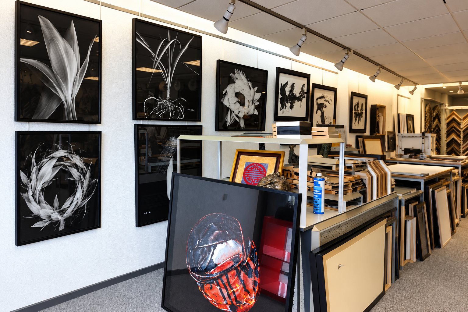Intérieur de l’atelier-galerie : tableaux encadrés de noir aux murs, rayonnages de baguettes, grand cadre posé au sol