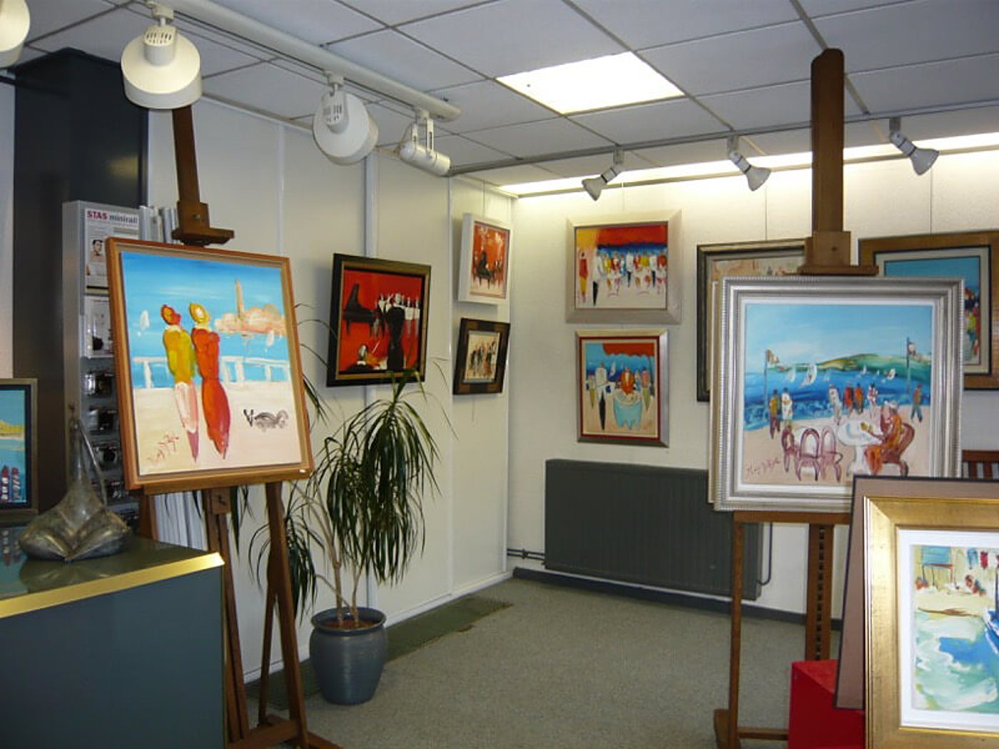 Intérieur d'une galerie d'art lumineuse où des tableaux colorés représentant des scènes de plage sont exposés sur des chevalets et accrochés aux murs.
