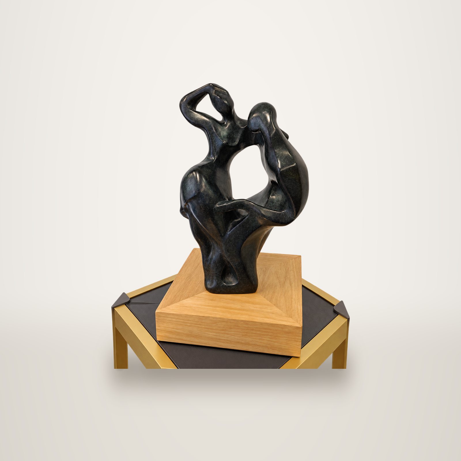 Sculpture abstraite en bronze noir représentant deux figures enlacées, posée sur un socle en chêne clair sur une petite table dorée devant un mur blanc.