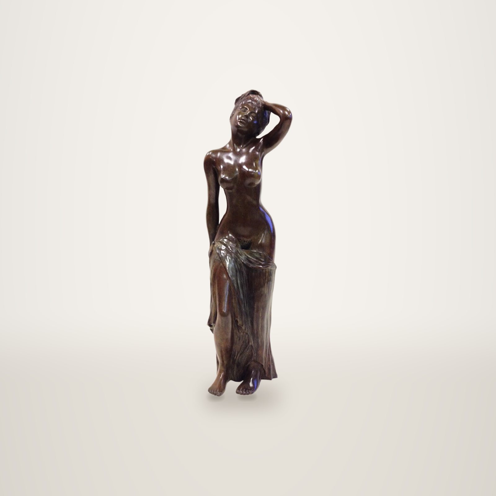 Statuette en bronze brun patiné représentant une femme debout, un bras relevé derrière la tête, une draperie nouée autour des hanches, posée sur une surface blanche devant un mur clair.